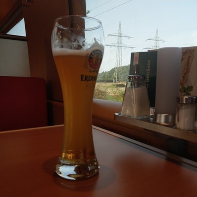 Ein fast volles Weissbierglas (konnte nicht bis zum foto warten) auf einem Tisch  in der Bahn vorm Fenster. Am Fenster Zucker, Licht, Salz, Pfeffer und Speisekarten. Vorm Fenster ein abgeerntetes Feld mit zwei Hochspannungsmasten