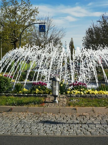 Ein Blumen gesäumter großer Springbrunnen in einem Kreisverkehr. Dahinter quert ein Seiltänzer die Straße (Kunnstobjekt)
