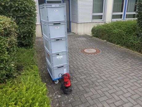 Fünf Euroboxen T-Shirts stehen gestapelt neben einer kleinen Tauchpumpe vor einem Gebäude.