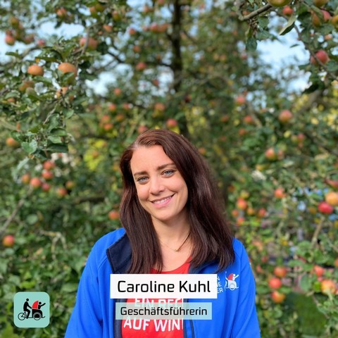Vor einem Apfelbaum steht eine Frau mit mittellangen, braunen Haaren in einer blauen Jacke und einem roten T-Shirt mit der Aufschrift "Jeder hat ein Recht auf Wind in den Haaren". Es ist Caroline Kuhl, die Geschäftsführerin von Radeln ohne Alter Deutschland. 