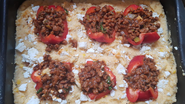 Eine rechteckige Metallform, darin gelbliche Polenta mit weißem Feta. Darauf liegen mit Veggie-Hack gefüllte Paprikaschiffchen. 