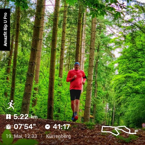 Foto von einem Läufer. Er trägt ein rotes Shirt, schwarze Radler und ein blaues Kopftuch. Er läuft auf einem Waldweg auf den Betrachter zu. Im Hintergrund sieht man Mischwald in strahlendem Grün.
