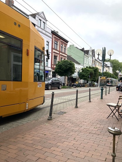 Straße mit Straßenbahnen, links kommt eine orangefarbene Straßenbahn ins Bild, rechts eine grüngemusterte. Im Hintergrund dazwischeb Altbauhäuser, vorne rote Strassenfliesen, 1 Bistrostuhl.