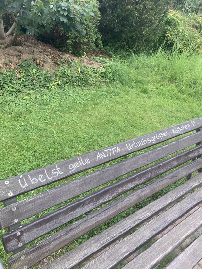 Auf einer Holzbank steht mit weißem Edding „Übelst geile Antifa Urlaubsgrüße! Küsschen!