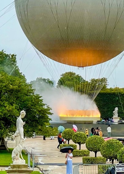 Heißluftballon in der Nähe eines Parks mit Menschen und Statuen, eine Person, die mit einem Regenschirm steht.