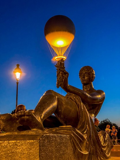 Eine Bronzestatue einer sitzenden Figur, die eine leuchtende Kugel hält, gegen einen tiefblauen Abendhimmel mit einer Straßenlaterne in der Nähe und Menschen im Hintergrund.