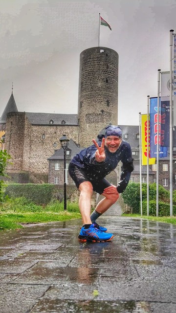 Das Bild zeigt eine Person, die ein Peace-Zeichen vor einem historischen Gebäude mit einem Turm macht. Es scheint regnerisch zu sein, da der Boden nass ist und die Person Regenkleidung trägt. Im Hintergrund sind Fahnen zu sehen. 
Am unteren Rand des Bildes sind Statistiken zu sehen: eine Distanz von 3,49 km, ein Tempo von 08'12