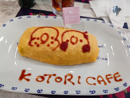 Ein Teller mit einer Portion Omrice (Ketchupreis umhüllt mit Omelett). Es ist mit Ketchup bemalt, mit einem Wellensittich- und einem Nymphensittichportrait. Unten drunter steht der Name des Cafés, das 