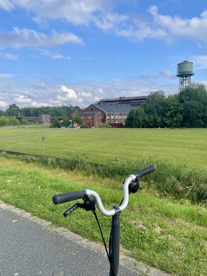 A brompton (foldable bike) handlebar is in the foreground with a grassy field, a few ducks, buildings of the Jahrhunderthalle, and a water tower in the background under a partly cloudy sky.

Ein Brompton-Lenker (faltbares Fahrrad) steht im Vordergrund mit einer gemähten Wiese, ein paar Enten, Gebäuden der Jahrhunderthalle und einem Wasserturm im Hintergrund unter einem leicht bewölkten Himmel bei gutem Wetter.