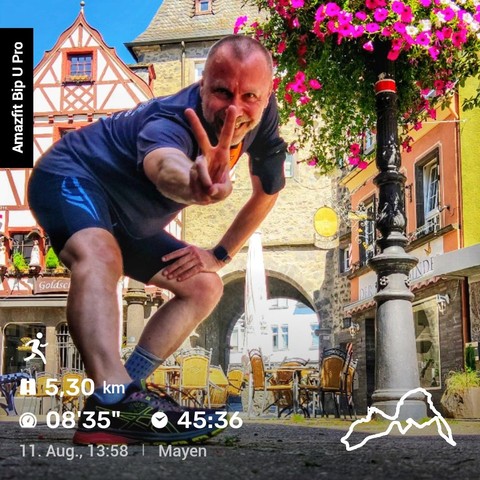 Das Bild zeigt eine fröhliche, sportliche Person in Sportkleidung, die sich vor einem malerischen Hintergrund in der Altstadt von Mayen, Deutschland, in die Kamera beugt. Der Mann trägt ein dunkles T-Shirt und Shorts, dazu Sportschuhe. Er macht ein Victory-Zeichen (Peace-Zeichen) mit der rechten Hand und lächelt in die Kamera.Im Hintergrund sind historische Fachwerkhäuser, eine Steinmauer und ein Torbogen zu sehen, die typisch für die Altstadtarchitektur sind. Auf der rechten Seite hängt ein Korb mit blühenden rosa Blumen, der dem Bild eine lebendige Note verleiht. Das Bild vermittelt eine positive und energiegeladene Stimmung.Am linken Rand des Bildes steht „Amazfit Bip U Pro“, was auf das verwendete Gerät zur Aufzeichnung der Laufdaten hinweist. Im unteren Bereich des Bildes sind die Laufstatistiken des Mannes abgebildet: eine Strecke von 5,30 km mit einer durchschnittlichen Pace von 08'35