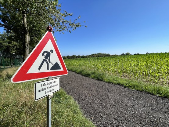 Warnschild für Straßenarbeiten neben einem Schotterweg in der Nähe eines Feldes und von Bäumen, unter einem klaren blauen Himmel. Schildtext: 