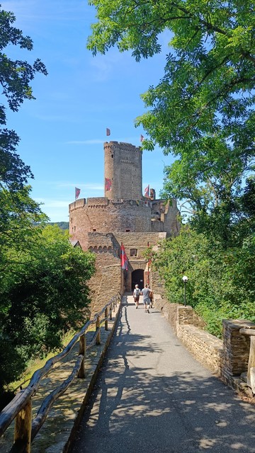 Blick auf die Ehrenburg an der Mosel. Eine mit Flaggen geschmückte Zugbrücke führt zum Tor, dahinter ragt die Runde Wehrburg und der Bergfried auf.