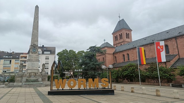 Blick auf einen grauen Obelisken links und eine rote Sandsteinkirche rechts. Davor steht ein goldener Schriftzug 'WORMS'