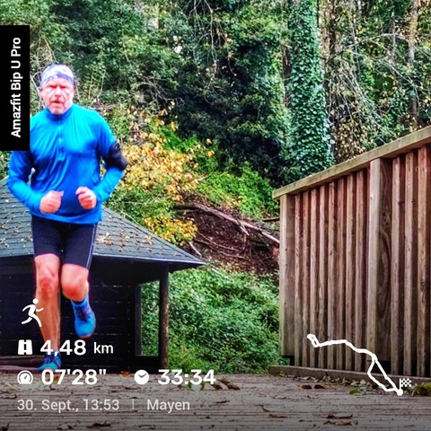 Das Bild zeigt eine Person, die in sportlicher Laufkleidung eine Strecke entlangläuft. Die Person trägt ein blaues Langarmshirt, eine schwarze Shorts und ein Stirnband. Im Hintergrund ist eine hölzerne Brücke oder ein Geländer zu sehen, sowie dichte grüne Vegetation und einen überdachten Holzunterstand. Auf der linken Seite des Bildes gibt es eine Anzeige mit der Marke 