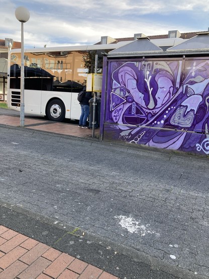 Im Vordergrund Busfahrstrasse des Busbahnhofs, dann rechts ein stehender Buss, halbiert durch das davor befindliche Bushäuschen mit einer Graffiti-Rückwand in violetten Farbtönen. 