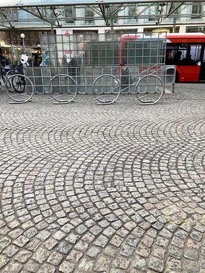 Im Vordergrund ein klassisches In Rundbögen verlegtes Kopfsteinpflaster . 4 kreisförmige Fahrradständer sind zu sehen, dahinter der Eingang zur Tiefgarage, dahinter ein Bushäuschen mit Sitzbank hinter Glas. 