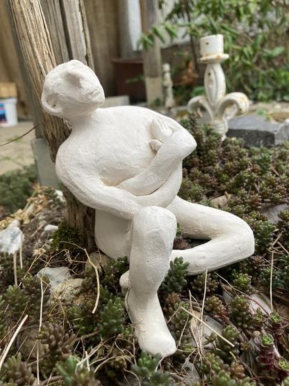 Sitzende männliche Figur aus weißem Ton nach dem Hochtemperatur-Schrühbrand, die ich gerade in ein Beet aus Sedum-Album/Mauerpfeffer gestellt habe. Sie lehnt an einem Clematis-Stamm.