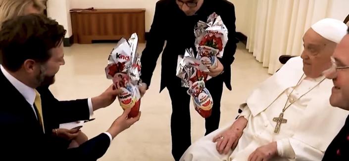 Rechts: Papst Franziskus in weißem Gewand und mit einem Kreuz um den Hals blickt freundlich auf die Menschen vor ihm. 
Links: J.D. Vance nimmt ein großes, in bunte Folie verpacktes Kinderschokoladen-Ei entgegen. 
Mitte: Ein weiterer Mann hält mehrere dieser Eier in den Händen.
Quelle: YouTube-Screenshot, The Australian