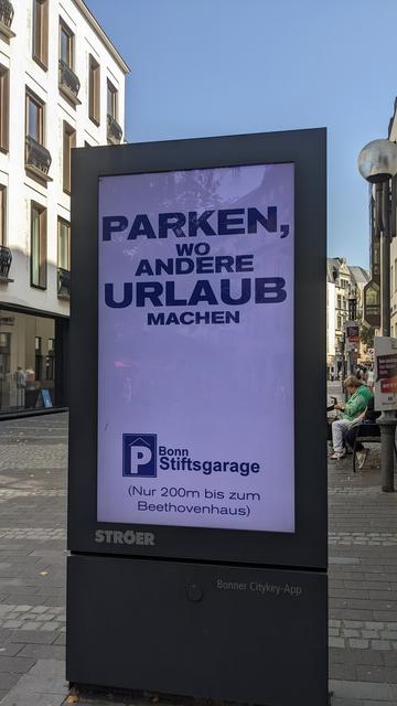 Eine digitale Werbetafel in der Fußgängerzone. Geworben wird mit "Parken, wo andere Urlaub machen" für die Stiftsgarage in Bonn.