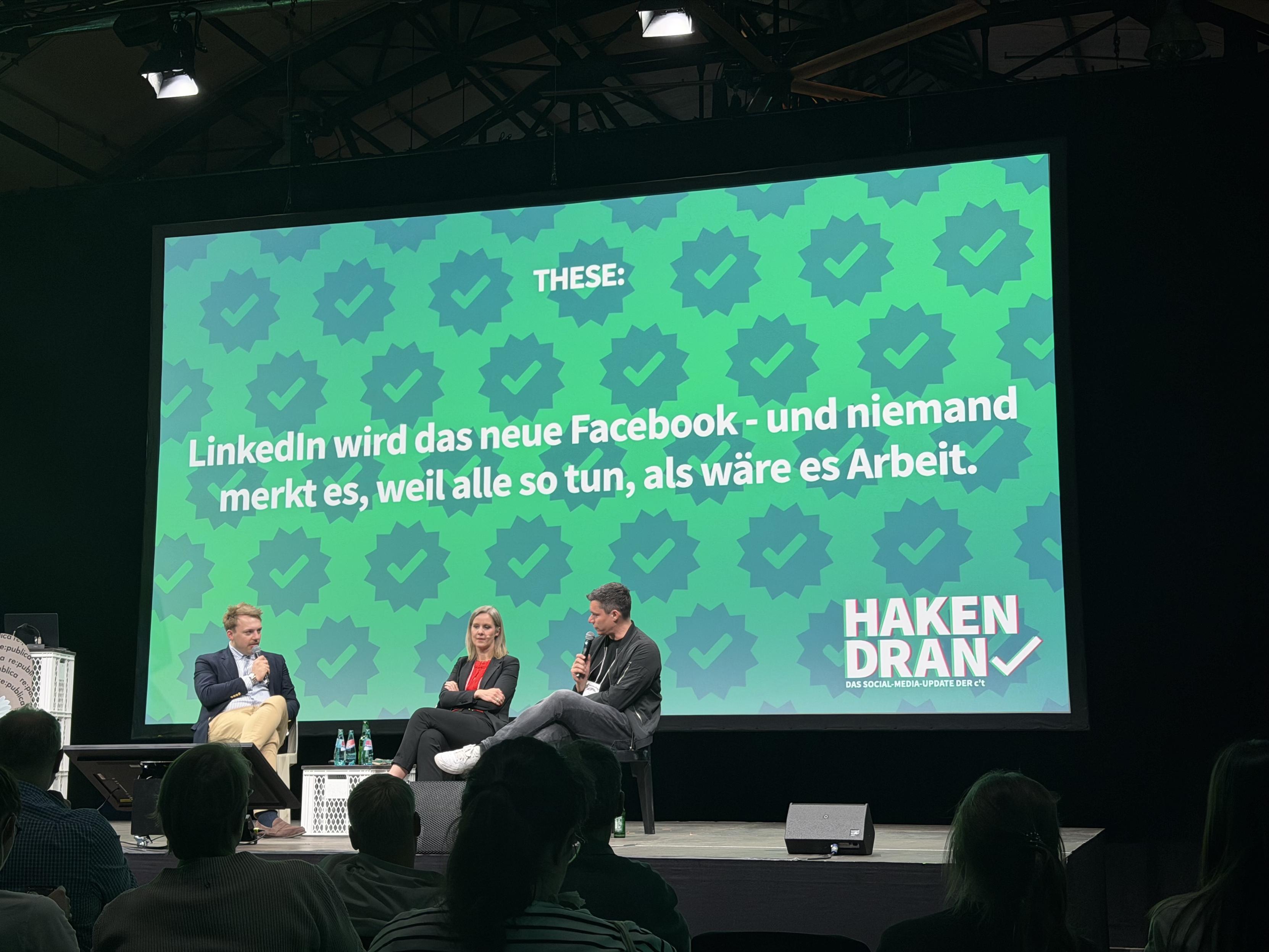 Auf der Bühne findet eine Podiumsdiskussion mit drei Referenten statt. Auf einer großen Leinwand im Hintergrund ist ein Text in deutscher Sprache zu sehen, der lautet: „LinkedIn wird das neue Facebook – und niemand merkt es, weil alle so tun, als wäre es Arbeit.“