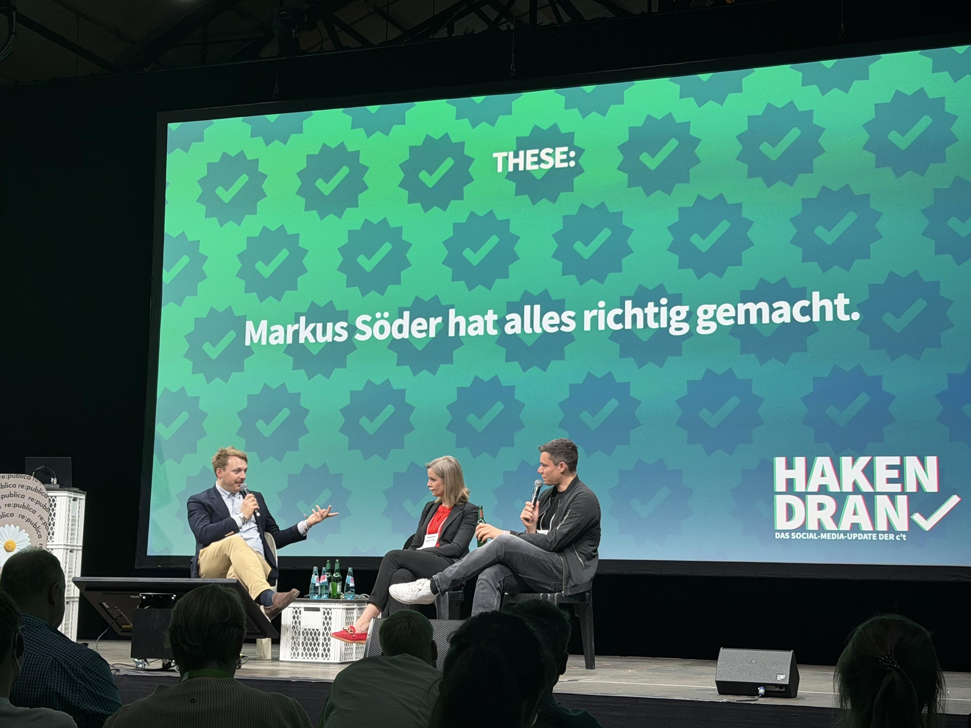 Auf der Bühne findet eine Podiumsdiskussion mit drei Referenten statt. Auf einer großen Leinwand im Hintergrund ist ein Text in deutscher Sprache zu sehen, der lautet: „Markus Söder hat alles richtig gemacht.“