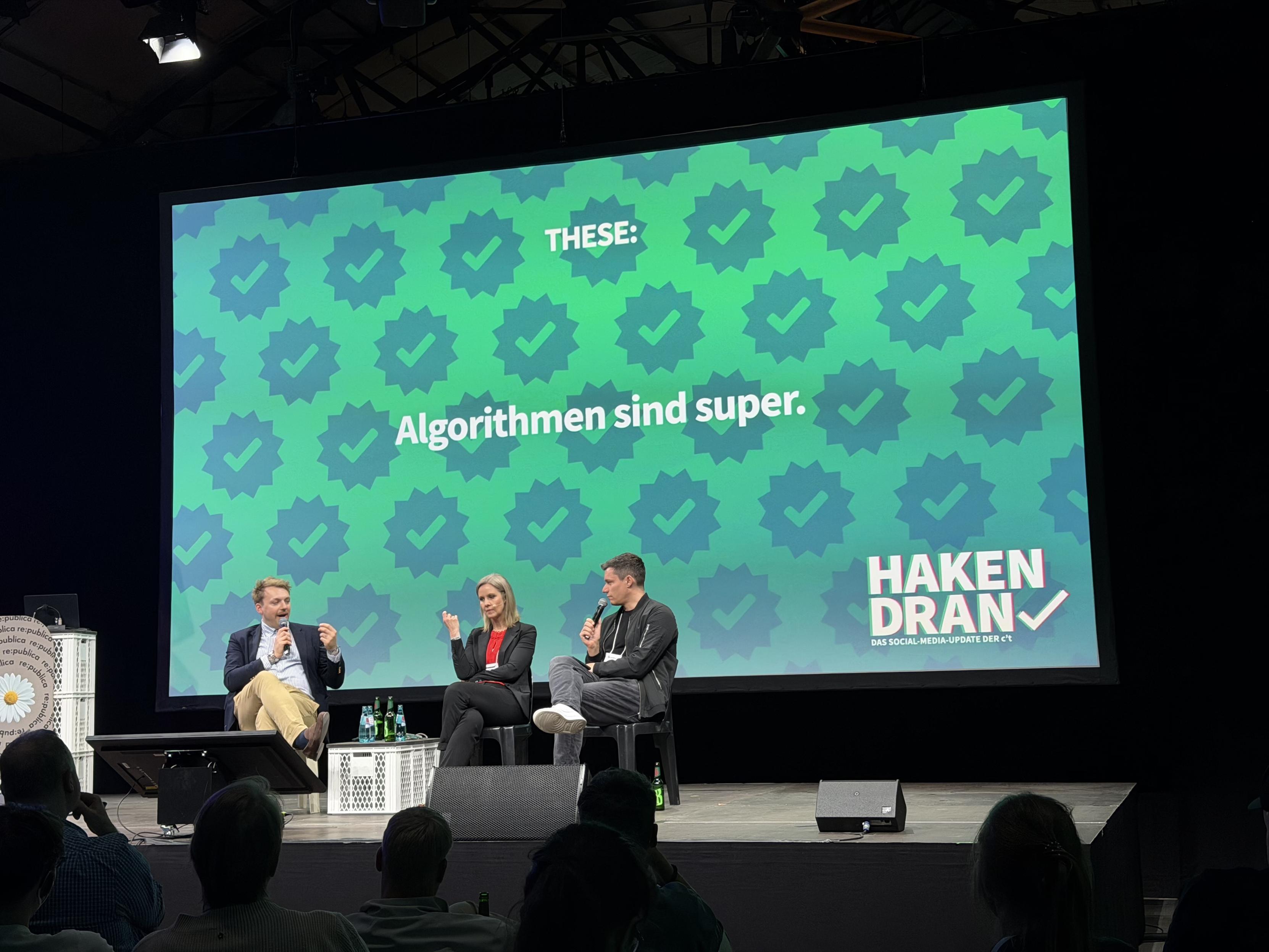 Auf der Bühne findet eine Podiumsdiskussion mit drei Referenten statt. Auf einer großen Leinwand im Hintergrund ist ein Text in deutscher Sprache zu sehen, der lautet: „Algorithmen sind super.“