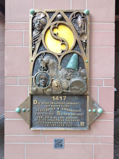 Bronzene Gedenktafel von Jürgen Goertz am Stadthaus am Dom auf rosa Fliesenwand. Links Relief mit Gesicht, Muschel- und Fischmotiven als Symbole der Sinti-und-Roma-Kultur. Inschrift: „Die Stadt erinnert mit dieser Tafel an den ersten urkundlichen Nachweis von Sinti in der Stadt Frankfurt am Main. Erwähnt wird ihre Ankunft in einem erhaltenen Dokument des Frankfurter Rates aus dem Jahr 1417.“ Darunter ein QR-Code.