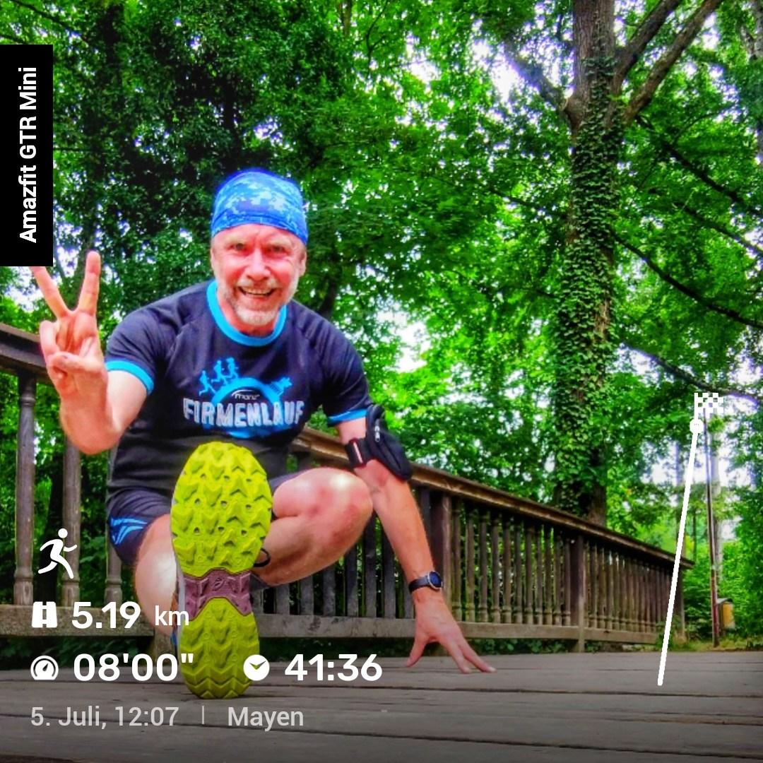 Eine Person steht auf einer Holzbrücke im Grünen, trägt ein schwarzes und blaues Laufshirt mit der Aufschrift ‚Firmenlauf‘, ein blaues Stirnband und Schuhe mit gelben Sohlen. Sie posiert mit angewinkeltem Knie und einem Peace‑Zeichen. Eingeblendet sind Laufdaten: 5,19 km, Pace 08:00 min/km, Dauer 41:36, aufgenommen am 5. Juli um 12:07 in Mayen.