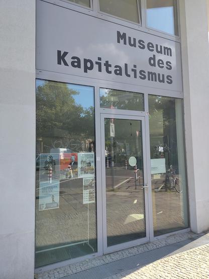 Gebäudeeingang mit Glastür und dem Schriftzug „Museum des Kapitalismus" über dem Eingang, davor ein Fahrrad und Bäume.