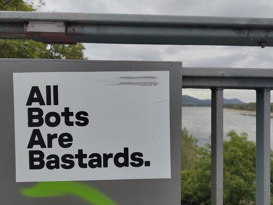 Schild mit der Aufschrift „All Bots Are Bastards“ an einem Metallgeländer, im Hintergrund ein Fluss mit bewaldeten Bergen unter bewölktem Himmel
