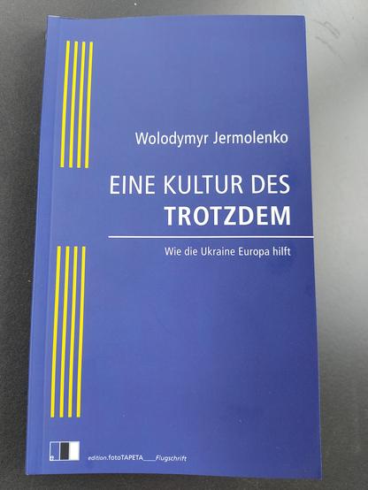 Buchcover mit blauem Grund: großer weißer Titel „EINE KULTUR DES TROTZDEM", darunter der Untertitel „Wie die Ukraine Europa hebt" und links der Autorenname „Wolodymyr Jermolenko". Unten drei gelbe Querstreifen sowie das Verlagslogo „edition.foto.to/TAPETA".